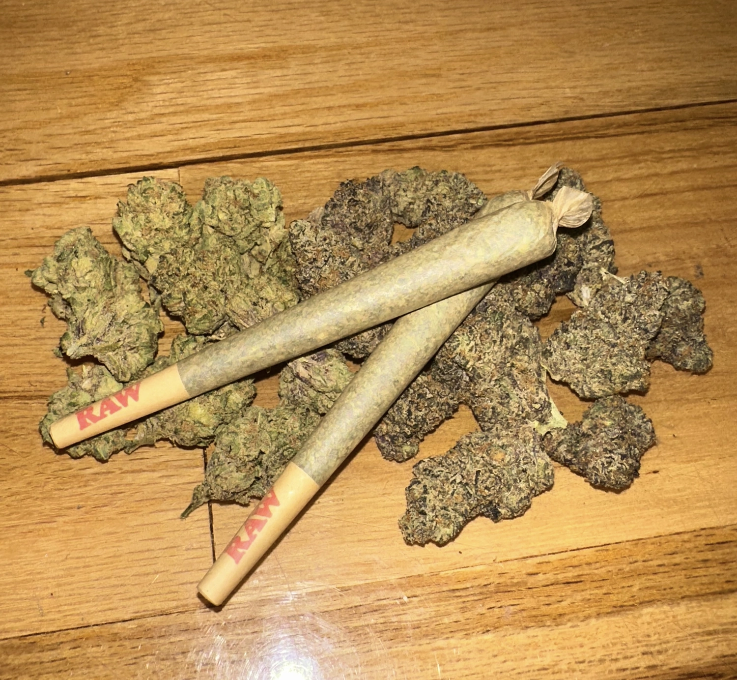 Two RAW brand pre-rolled joints positioned atop various cannabis buds on a wooden surface.