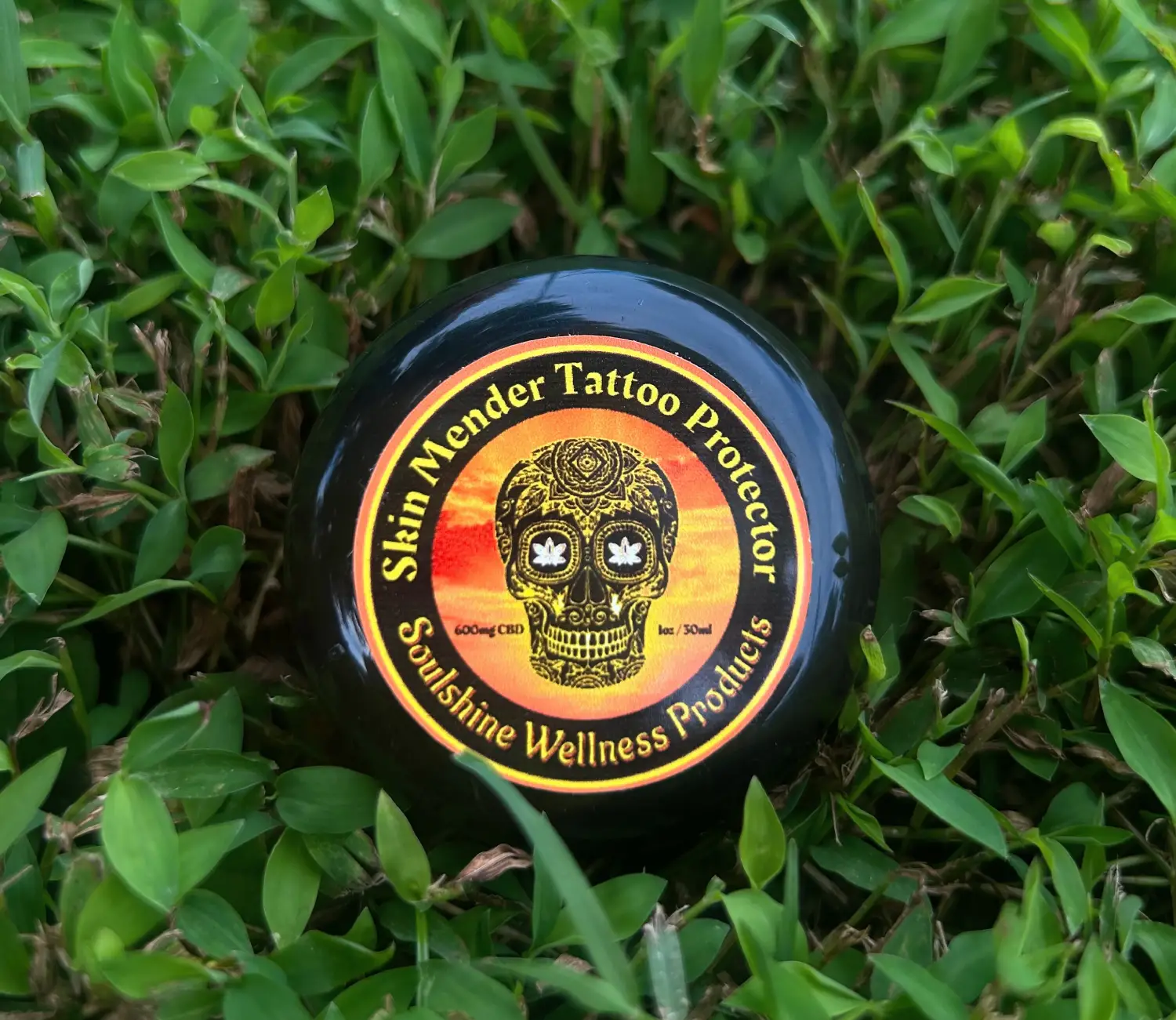 A black jar of Skin Mender Tattoo Protector placed on lush green grass, labeled as a product by Soulshine Wellness.
